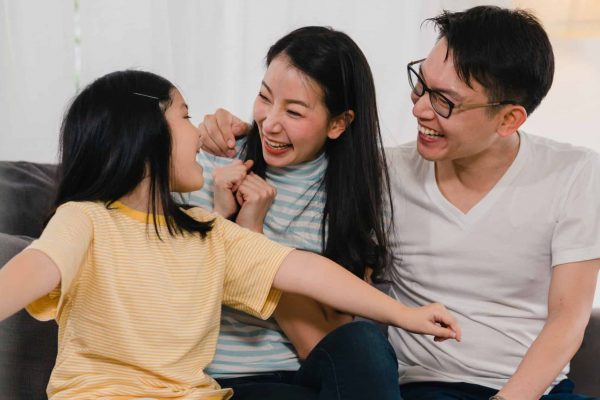 Happy Asian young family homeowners bought new house. Japanese Mom, Dad, and daughter embracing looking forward to future in new home after moving in relocation sitting on sofa with boxes together.